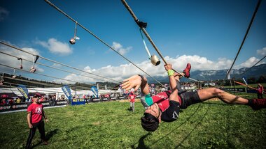 Participant during a Spartan Race climbing on ropes obstacle on a grassy field with spectators in the background. | © Michael Vorbrüggen