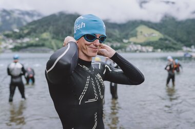 Person in wetsuit with swimming cap and goggles preparing for water sports in a lake, with mountains and clouds in the background. | © EXPA, Jürgen Feichter