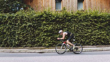 Person cycling on the street in front of a large green bush and a wooden house. | © EXPA, Jürgen Feichter