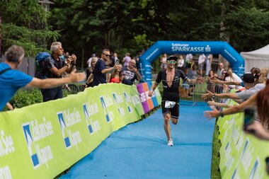 A runner crosses the finish line at an outdoor race, surrounded by cheering spectators and a blue finish arch. | © Zell am See-Kaprun Tourismus