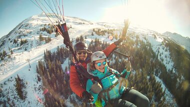 Two paragliders flying in the mountains with snow-covered peaks and a forest illuminated by sunlight. | © FalkenAir Tandemparagliding