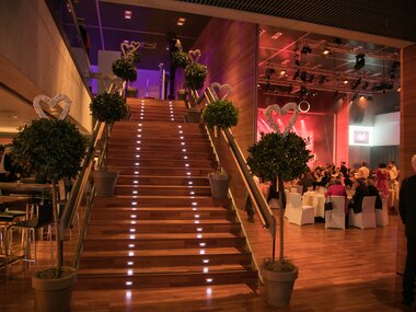 Interior of a festive decorated event space with an illuminated staircase, heart decorations, and tables in the background. | © Nikolaus Faistauer Photography