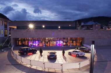 Exterior view of the entrance area of a modern building at night, with parked cars and colorful lights inside. | © FPCC