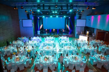 Elegant event space with multiple round tables set for a formal evening reception, illuminated with blue lighting. | © Nikolaus Faistauer Photography