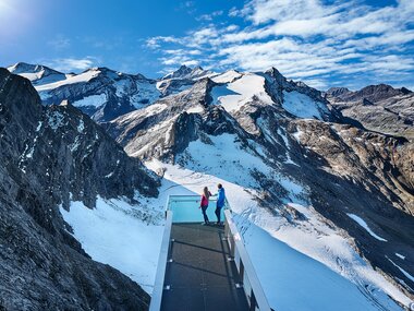 Two people stand on a glass bridge in the Alps, surrounded by snow-covered peaks and blue sky. | © Kitzsteinhorn 