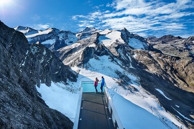 Two people stand on a glass bridge in the Alps, surrounded by snow-covered peaks and blue sky. | © Kitzsteinhorn 
