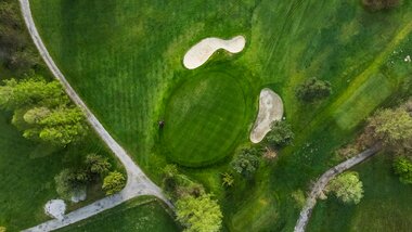 Aerial view of a golf course featuring a lush green fairway, bunkers, and pathways surrounded by trees. | © Zell am See-Kaprun Tourismus