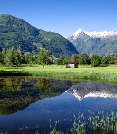 Landscape with a pond surrounded by trees, mountains in the background, and a clear blue sky. | © Golfclub Zell am See