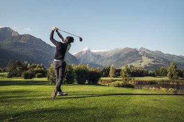 Person playing golf on a green field with mountains and a lake in the background on a sunny day. | © Zell am See-Kaprun Tourismus
