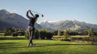 Person playing golf on a green field with mountains and a lake in the background on a sunny day. | © Zell am See-Kaprun Tourismus