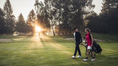Two people playing golf on a well-maintained golf course at sunrise, with trees in the background. | © Zell am See-Kaprun Tourismus