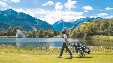 Golfer pulls their equipment across a lush golf course with mountain scenery in the background. | © Johannes Radlwimmer 