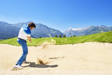 A golfer hits the ball out of a bunker on a golf course with mountain views. | © Golfclub Zell am See
