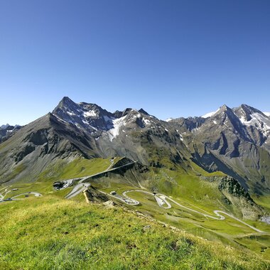 View of alpine landscape with winding road and tall mountains, snow on peaks, under a clear blue sky. | © Großglockner Hochalpenstraße 