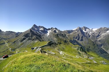 View of alpine landscape with winding road and tall mountains, snow on peaks, under a clear blue sky. | © Großglockner Hochalpenstraße 