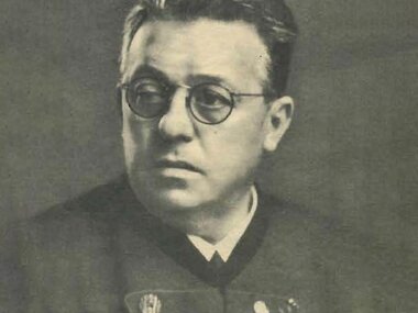 Black-and-white photo of a person with glasses, wearing a dark jacket with badges.