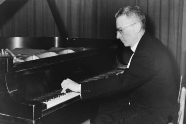 Black and white photo of a person sitting at and playing a piano with a serious expression.