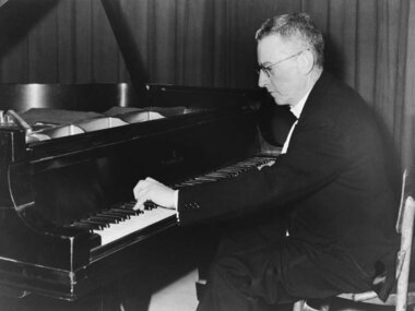 Black and white photo of a person sitting at and playing a piano with a serious expression.
