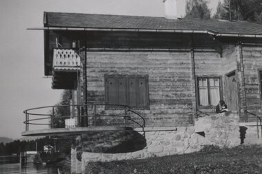 Black and white photo of a wooden house by the water with a balcony, likely by a lake, with a person sitting on the terrace. | © Irmgard Blaickner