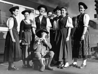 Black and white photo of the Trapp family in traditional costumes in front of a train, likely from the mid-20th century. | © www.photopress.at