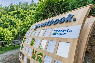 Wooden guestbook with various lists and texts posted, along a trail in lush greenery under a blue sky.