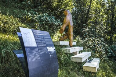 Outdoor sculpture on a staircase with an information plaque, surrounded by trees and nature.