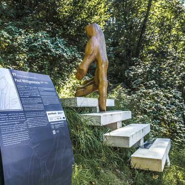 Outdoor sculpture on a staircase with an information plaque, surrounded by trees and nature.