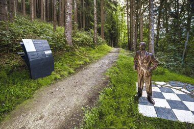 Sculpture in the forest featuring a path, an informational sign, and a metal figure on a black-and-white checkered pattern