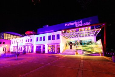 The indoor swimming pool Halle is in a modern building illuminated with colorful lighting at night. | © Freges