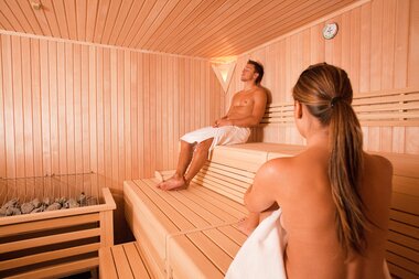 Two individuals relaxing in a sauna, one sitting on an elevated wooden bench, the other sitting below, both with towels. | © Johannes Felsch