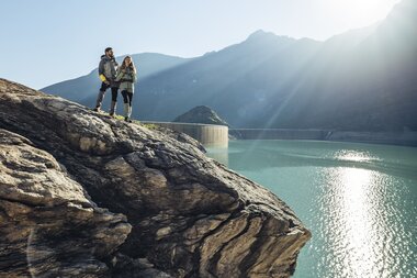 Two people are standing on a large rock by a high mountain lake with mountains in the background during sunrise or sunset. | © Zell am See-Kaprun Tourismus