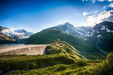 High mountain reservoirs with water, surrounded by tall snow-capped peaks and green slopes, under a sunny sky. | © Christian Mairitsch