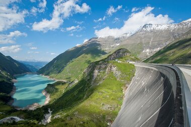 High mountain reservoir with a dam, surrounded by green mountains under a blue sky. | © VERBUND 
