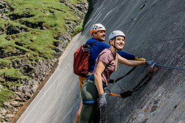 Two climbers with gear ascending a steep concrete wall in high mountain terrain. | © Zell am See-Kaprun Tourismus