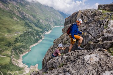 Two climbers on a rocky wall overlooking a large alpine lake and green mountains. | © Zell am See-Kaprun Tourismus