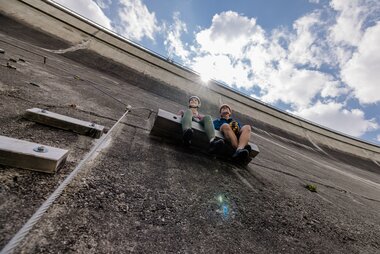 Two climbers resting on a ladder on a steep wall with clouds in the sky above. | © Zell am See-Kaprun Tourismus