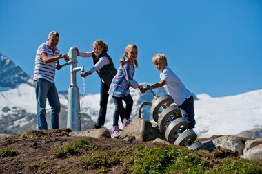 People drawing water from a mountain spring with snowy peaks in the background. | © VERBUND 