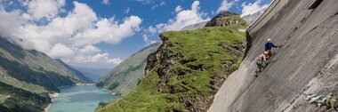Climbers on a cable bridge on a steep slope with a lake and mountains in the background. | © Zell am See-Kaprun Tourismus