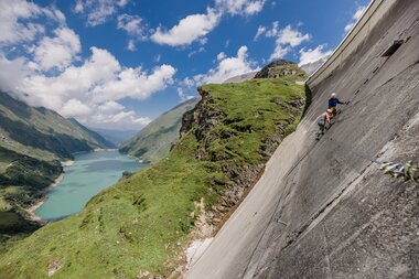 Climbers on a cable bridge on a steep slope with a lake and mountains in the background. | © Zell am See-Kaprun Tourismus