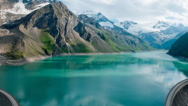 View of a large high mountain reservoir surrounded by snow-capped mountains and green slopes. | © VERBUND