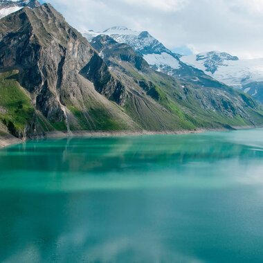 View of a large high mountain reservoir surrounded by snow-capped mountains and green slopes. | © VERBUND
