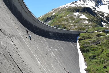 High mountain reservoir with a large dam, mountains in the background, and blue sky. | © VERBUND