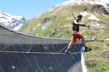 Person climbing a high mountain dam wall under a blue sky with mountain scenery. | © VERBUND