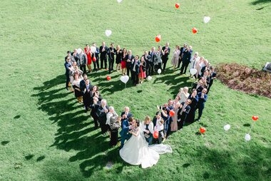Wedding guests forming a heart shape on green lawn, with balloons floating in the air. | © Claire Morgan 