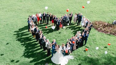 Wedding guests forming a heart shape on green lawn, with balloons floating in the air. | © Claire Morgan 