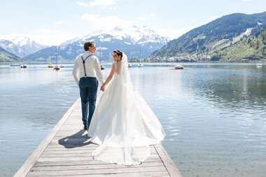 A wedding couple holding hands on a dock at a lake with mountains in the background on a clear day. | © Schloss Prielau 