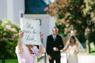 Wedding scene with a person holding a sign that says 'Here comes the bride,' in front of a church-like building and park. | © Claire Morgan