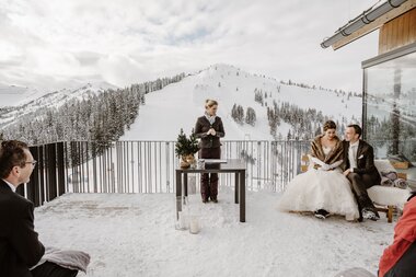 Wedding ceremony in a snowy mountain village with a view of snow-covered mountains and a group of people in formal attire. | © Wild Connections Photography