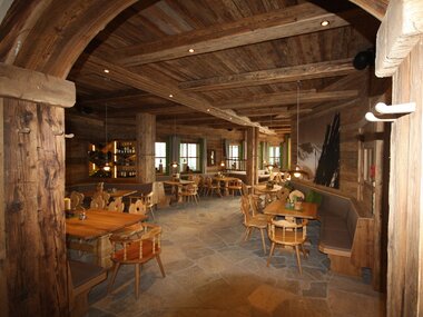 Interior of a rustic hotel with wooden beams, cozy tables and chairs, natural light through windows. | © Berghotel Schmittenhöhe
