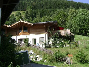 Traditional wooden house in nature with a terrace, surrounded by trees and hills, on a sunny day. | © Augut Zell am See